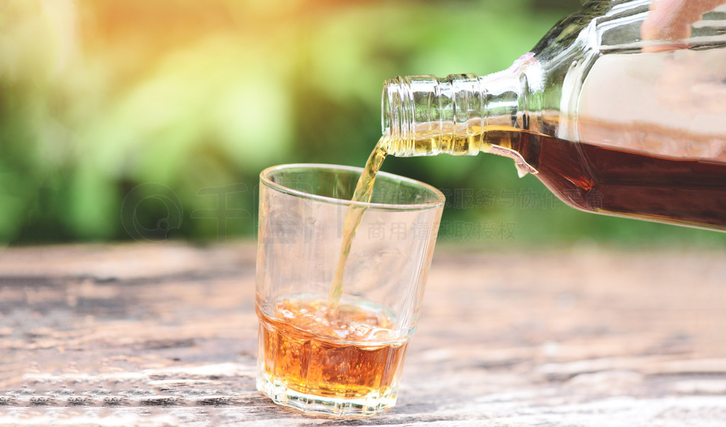 在木质背景上将威士忌或酒精饮料从瓶子倒入玻璃杯/倒酒